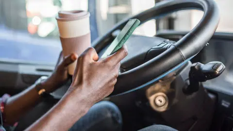 Getty Images A man sits in a stationary vehicle in front of a steering wheel holding a cup of coffee in one hand and a phone in the other