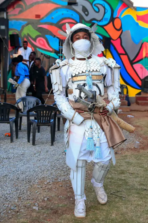 PHILIMON BULAWAYO / REUTERS Cosplay player Gabriel Ota walks during an anime, manga and cosplay meeting. He is dressed as popular Japanese action game character, Jin Sakai, the sword-wielding protagonist of Ghost of Tsushima. He is wearing a white samurai outfit made of cardboard and recycled paper.