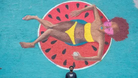  Krisztian Elek / Getty Images An image of South African artist Breeze Yoko standing in front of a mural of a woman wearing a yellow bathing suit, sitting in a float in the shape of a watermelon on 31 July 2024.