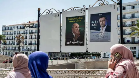 AFP Women walk past a notice board depicting posters for presidential election candidates incumbent President Abdelmajid Tebboune (L) and the Socialist Forces Front's Youcef Aouchiche, in the centre of Algiers - 5 September 2024