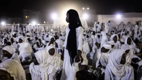 John Wessels/AFP  An image of a young woman standing in front of bright white light creating a halo-like effect, surrounded by other women who are sitting down and dressed in fully white garments in Dakar, Senegal - Sunday 15 September 2024.