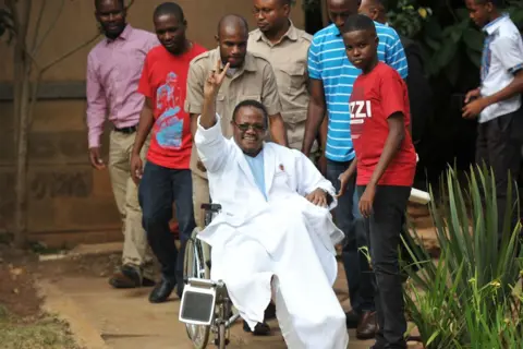 AFP Tundu Lissu waves from his wheelchair after giving a press conference surrounded by members of his family and supporters on 5 January 2018 at a hospital in the Kenyan capital, Nairobi