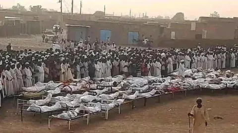 X A large group of mourners stand in an open field beside a number of bodies wrapped in cloth.