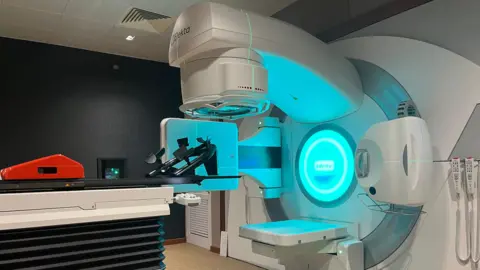 A radiotherapy machine in a hospital lit up with blue light. There is a bed for patients to lie on with a headrest.