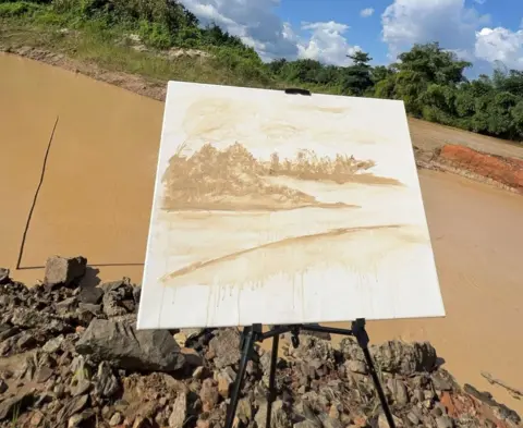 Jay Sterling A painting of the Pra River using a yellow-brown colour on a white canvas, on a stand in front of the polluted river