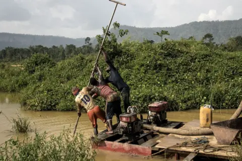 Getty Images Three Illegal gold panners stand on a motorised pump to scrape the river bed as they look for specks of gold in the Kibi area in southern Ghana on 12 April 2017. The river is yellow-brown, with thick bushes on either side.