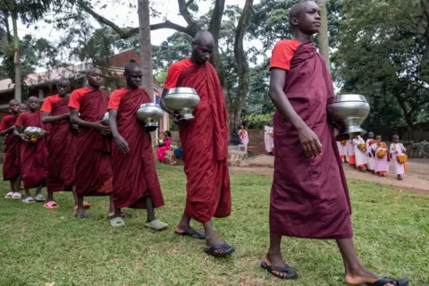 BADRU KATUMBA/AFP Ugandan novice monks dressed in shades of red and brown are queuing up to receive an offering from the community after meditation celebrating Kathina, a significant event on the Buddhist calendar in Entebbe, on 20 October. Bhante Buddharakkhita, formerly known as Steven Jemba Kabogozza, brought Buddhism to Uganda 20 years ago after returning from India. Since 2005, he has been teaching mindfulness meditation across Africa. His ambitious goal is to train at least 54 monks and send each one to a different African country to teach and spread the message of Buddhism within the context of the African culture.