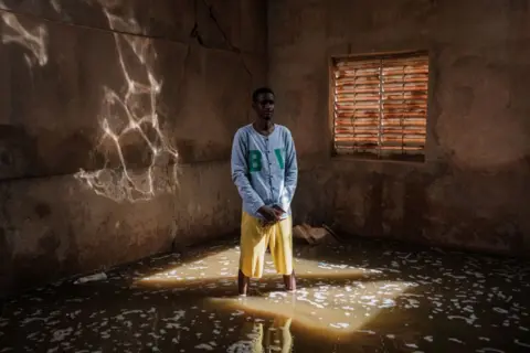 GUY PETERSON/AFP Teacher Suleyman Ba stands in a classroom of the Ecole de Diogel with water marks from flooding less then a week before and water still ankle deep in class rooms and waste deep in the court yard in Dioguel on October 22, 2024. Floods along the Senegal river have affected over 55,000 people after heavy rain in the Senegal River Basin leaving many villages underwater and over 1,000 hectares of farm land submerged. 