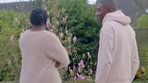 Fatoumata Sylla (l) looking out over a flowering garden with her arms crossed and her back to the camera. Her brother Abdoul (r) is looking at her - in Paris, France. Both are wearing salmon-pink tops