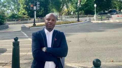 Yves Kaduli Dr Yves Kaduli, a 38-year-old asylum seeker from the Democratic Republic of Congo, stands in front of the Capitol Building with his arms folded in a suit jacket 