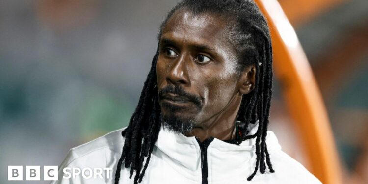 A head shot of Aliou Cisse wearing a white tracksuit top with his dreadlocks hanging loose