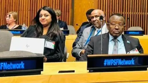 Somalia's Family Ministry / X Somalia’s Family Minister Gen Bashir Mohamed Jama pictured in a conference room with other delegates, including Somali politician Abdullahi Godah Barre who is looking over his shoulder into the camera. A female delegate with long hair is seated next to them both and is also looking into the camera - New York, US - 22 October 2024