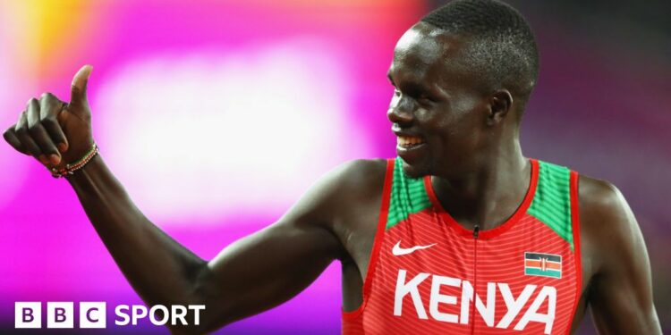 Kipyegon Bett, wearing a red and green athletics vest, gives a 'thumbs up' gesture while looking to his right during a track meeting