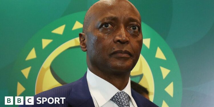 A headshot of Patrice Motsepe wearing tie and suit standing in front of a Confederation of African Football banner