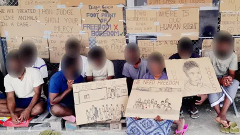 BBC Tamil migrants on Diego Garcia, with their faces blurred, holding protest signs reading 