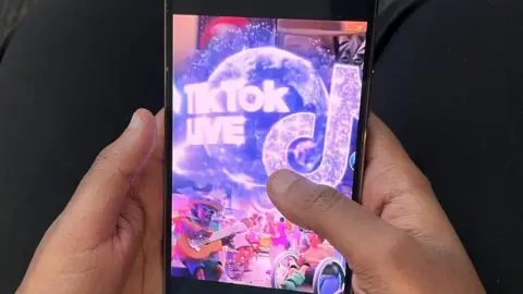 BBC Shows someone's hands holding a mobile phone with TikTok Live on the screen.