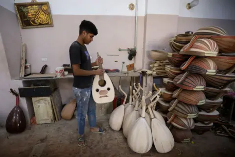 Ahmad Hasaballah / Getty Images  Young man holding Traditional Oud Instrument at workshop belonging to Khaled Azzouz, a longtime maker of the traditional oud instrument. The instruments are piled up in rows next to a wall.