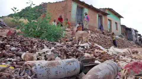 Amensisa Negera / BBC Shells and other rusting ammunition lie on the ground in front of a few houses, some of whose inhabitants are seen sitting outside. A few stones have been painted red as a warning and a goat is nearby. Tigray, Ethiopia - October 2024