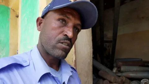 Amensisa Negera / BBC Police commander Hadush Gebremedhin in his blue uniform seen with a room full of weapons like mortar shells behind him 