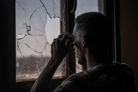 Amaury Falt-Brown / AFP A bearded army soldier, viewed from behind, scans the frontline with binoculars through a broken window in Khartoum, Sudan - Sunday 3 November 2024