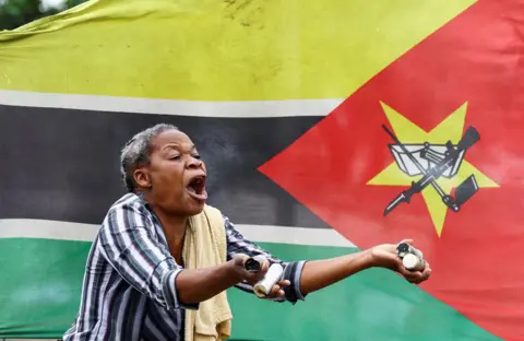 Siphiwe Sibeko / Reuters A woman in a stripped shirt with a towel over her shoulder shouts and holds up empty tear gas cannisters at a protest in Maputo. Behind her is the Mozambican flag - Thursday 7 November 2024