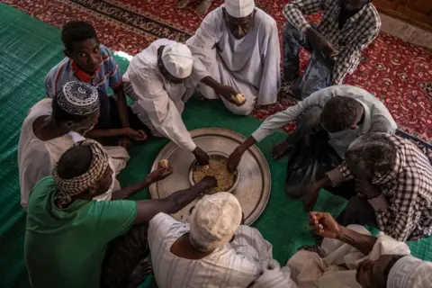 Amaury Falt-Brown / AFP Men sit on the floor covered with mats around a dish placed on a silver platter. They  are sharing the food, eating with their hands, in Omdurman, Sudan - Friday 1 November 2024