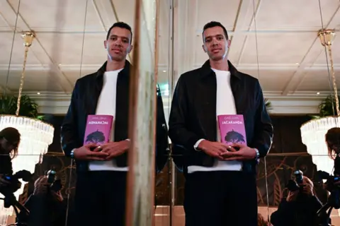 Julien de Rosa / AFP Rwandan-French writer and singer Gael Faye, whose image is also reflected in a mirror, poses with his book Jacaranda in Paris, France - Monday 4 November 2024