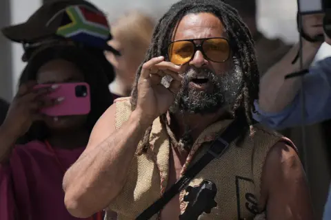 Nic Bothma / Reuters A protester with dreads, a beard and wearing orange lensed sunglasses shouts at Kalk Bay Harbour in Cape Town, South Africa - Thursday 7 November 2024 