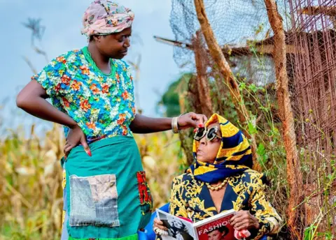 Luxury Media Zambia Diana Kaumba standing outside in traditional Zambian clothing looking down at her seated grandmother in patterned navy and gold designer jacket, scarf and sunglasses - holding a book with the title Hollywood Fashion, with Audrey Hepburn on the cover