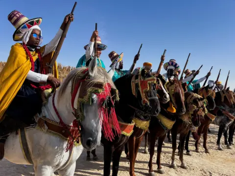 Abdelaziz Boumzar / Reuters A line of men on horse point their rifles to the sky as they await to start their performance at the International Festival of Saharan Tourism in Oued Souf, Algeria - Saturday 16 November 2024