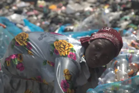 Abiba Alhassan, a woman wearing a floral dress and a headscarf, sorts out used plastic bottles next to the Agbogbloshie dumpsite in Accra