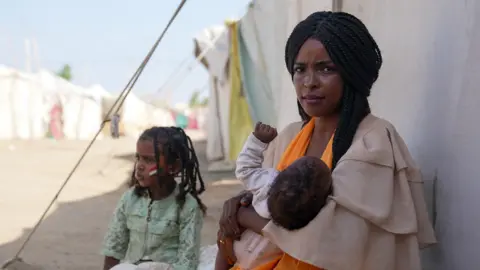 Joyce Liu / BBC A woman called Omnia sits outside a tent in Sudan holding her baby, with a second child sat next to her