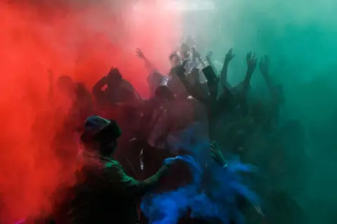 Gerald Anderson / Getty Images Crowds of smiling people are covered in multicoloured powder.