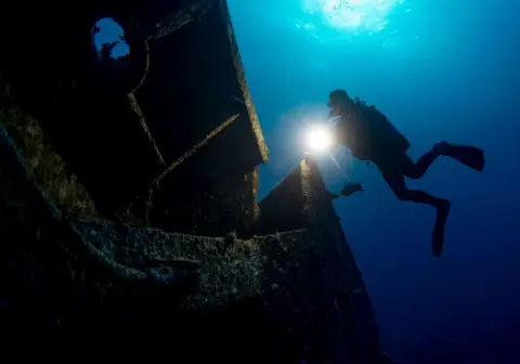 Mahmut Serdar Alakus / Getty Images A diver shines a flashlight under water at a wreck.