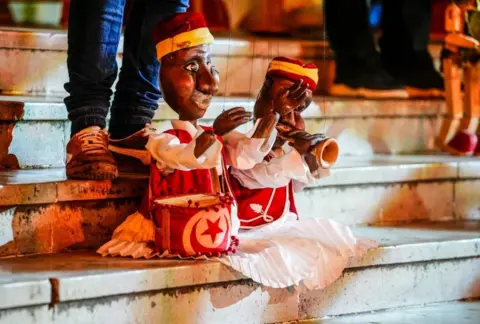 Yassine Gaidi / Getty Images A man manipulates two puppets on strings. The puppets are holding a drum and a trumpet.