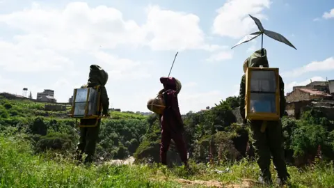 Kairos Futura Three people dressed in green suits and hoods walk in a green space.