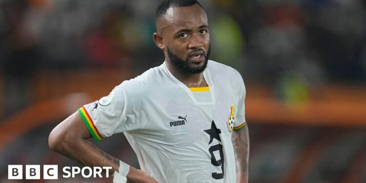 Jordan Ayew, wearing a white Ghana shirt with a number nine on the chest, stands with his hands on his hips