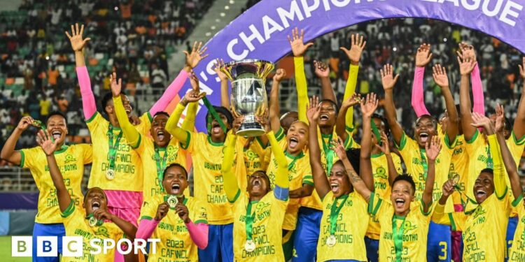 Mamelodi Sundowns players, all wearing matching yellow t-shirts and with gold medals around their necks, stand with their arms raised on a podium as  captain Zanele Nhlapho lifts the Caf Women's Champions League trophy