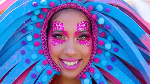 Chris Jackson / Getty Images A female performer smiles in a pink and blue headdress - with pink sequins around her eyes and pink lipstick poses at a carnival in Cape Town ahead of the 2024 Earthshot Prize - Wednesday 6 November 2024