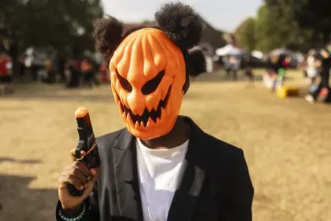 Kim Ludbrook / EPA A young fancy dress contestant - wearing a pumpkin mask, toy gun, black blazer and afro puffs - waits to take part in in the best-dressed competition during the annual Halloween party at the George Hay Park. The event aims to raise funds to maintain and improve the neighborhood's green spaces.