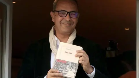 Getty Images A man, Kamel Daoud, in black-framed glasses smiles as he stands in an open window, holding a white copy of his prize-winning book. He is wearing a dark blazer and a white shirt, with a cream-coloured scarf lightly looped around his neck.