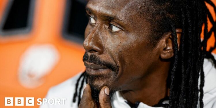 Aliou Cisse, with his back hair braided and a goatee beard, holds his left hand to his chin as he looks to his right