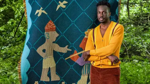 Alexander Edwards / Amechi Mandi Cameroonian-Nigerian interior designer Amechi Mandi, dressed in a yellow cardigan and burnt brown cords, stands in a garden with arms folded in front of one of his throw blankets called 'Chief Palaver' showing two figures and two birds stitched on to blue diamond designed thick woven fabric 