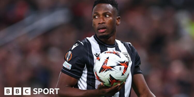 Baba Rahman, wearing a black and white striped PAOK shirt, holds a football in the palm of his right hand as he turns his head to his right over