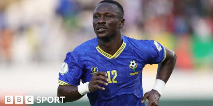 Simon Msuva, wearing a blue Tanzania shirt with yellow trim and the number 12 on the front, runs towards the camera during a football game