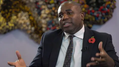 BBC David Lammy in a suit with a red Remembrance poppy on his lapel. He is talking and moving his hands as he talks.