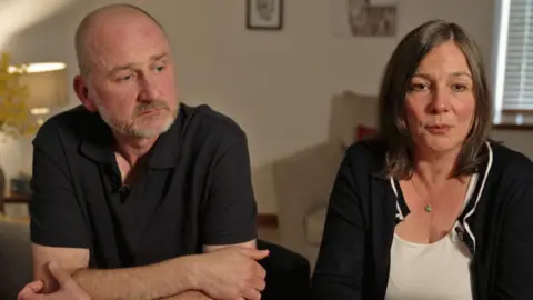 BBC Mark Dowey in a black polo shirt leans on a table looking off to the side in thought, beside his wife Ros in a white top and black cardigan who is looking into the camera. They sit in a light lounge with a lamp on in the background and the blinds drawn.