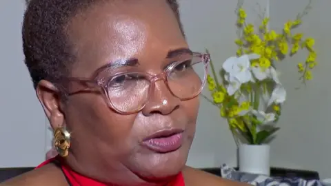 BBC Edith Nyachuru, seated and wearing a red top, reading glasses and gold earrings. A vase of flowers can be seen behind her - 13 November, Zimbabwe