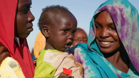 Joyce Liu / BBC A woman with a green and purple headscarf smiles at a baby that she is holding.