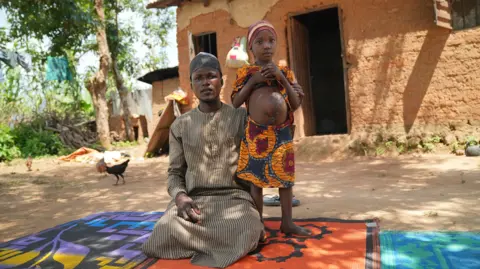 Gift Ufuoma / BBC Masud Abdulrasheed is sitting while his daughter Zaharau is standing next to him, showing her wound